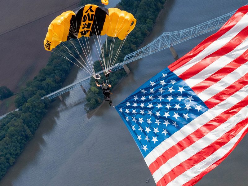 Army parachutist descending over a river beside a large American flag
